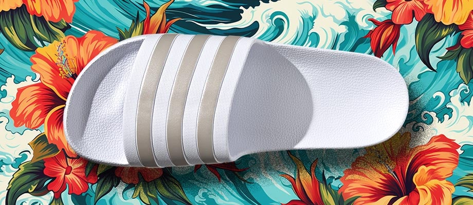 adidas aqua Adilette Badelatsche in Wei&szlig; mit goldbeigen Streifen von oben mit Blick auf den Riemen und das ergonomisch leicht geformte Fu&szlig;bett vor einem blumigen Hawaii-Hemden-Hintergrund mit Wellen, um den Namen Aqua und das Wasserthema zu unterstreichen