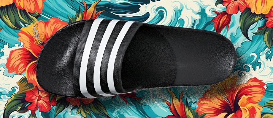adidas aqua Adilette Badelatsche in Shwarz von oben mit Blick auf den Riemen und das ergonomisch leicht geformte Fußbett vor einem blumigen Hawaii-Hemden-Hintergrund mit Wellen, um den Namen Aqua und das Wasserthema zu unterstreichen
