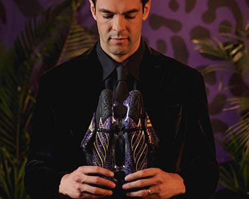 Kak&aacute; en costume noir devant un papier peint violet &agrave; motif jaguar avec des plantes en d&eacute;coration regardant sa paire de Predator signature avec design jaguar et accents dor&eacute;s