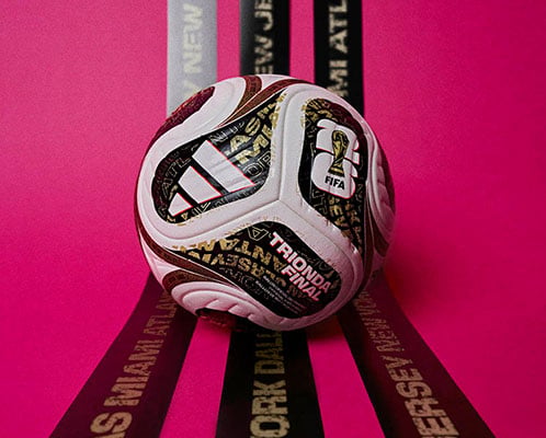 adidas WM 26 Final OMB Spielball vor pinkfarbenem Hinterdrund auf drei schwarzen Banderolen, die im Goldton der WM-Troph&auml;e die drei Finalaustragungsorte tragen
