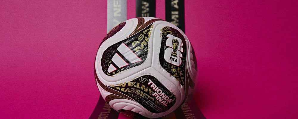 adidas WM 26 Final OMB Spielball vor pinkfarbenem Hinterdrund auf drei schwarzen Banderolen, die im Goldton der WM-Troph&auml;e die drei Finalaustragungsorte tragen