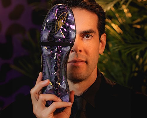 Gros plan sur le visage de Kak&aacute; devant un papier peint violet &agrave; motif jaguar avec des plantes en d&eacute;coration couvrant son &oelig;il droit avec sa chaussure signature face semelle montrant les crampons et le design de semelle avec sa signature en dor&eacute;